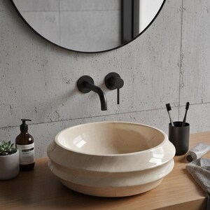May include: A cream-colored, layered ceramic sink with a black faucet and fixtures. The sink sits on a wooden countertop, with a round black mirror above. A soap dispenser, plant, and toothbrush holder are also visible.