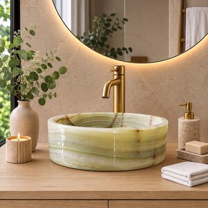May include: A round, light green onyx stone vessel sink with natural banding, paired with a brushed gold faucet. The sink sits on a light wood vanity alongside a beige ceramic vase with greenery, a lit candle, a soap dispenser, and a bar of soap.