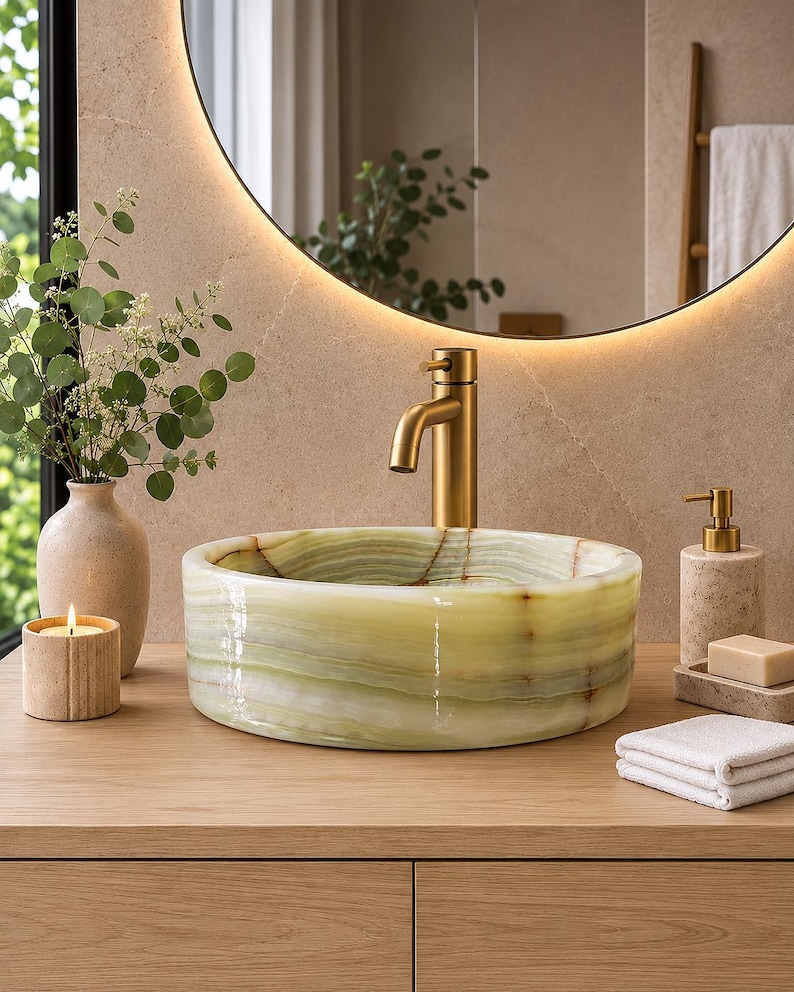 May include: A round, light green onyx stone vessel sink with natural banding, paired with a brushed gold faucet. The sink sits on a light wood vanity alongside a beige ceramic vase with greenery, a lit candle, a soap dispenser, and a bar of soap.