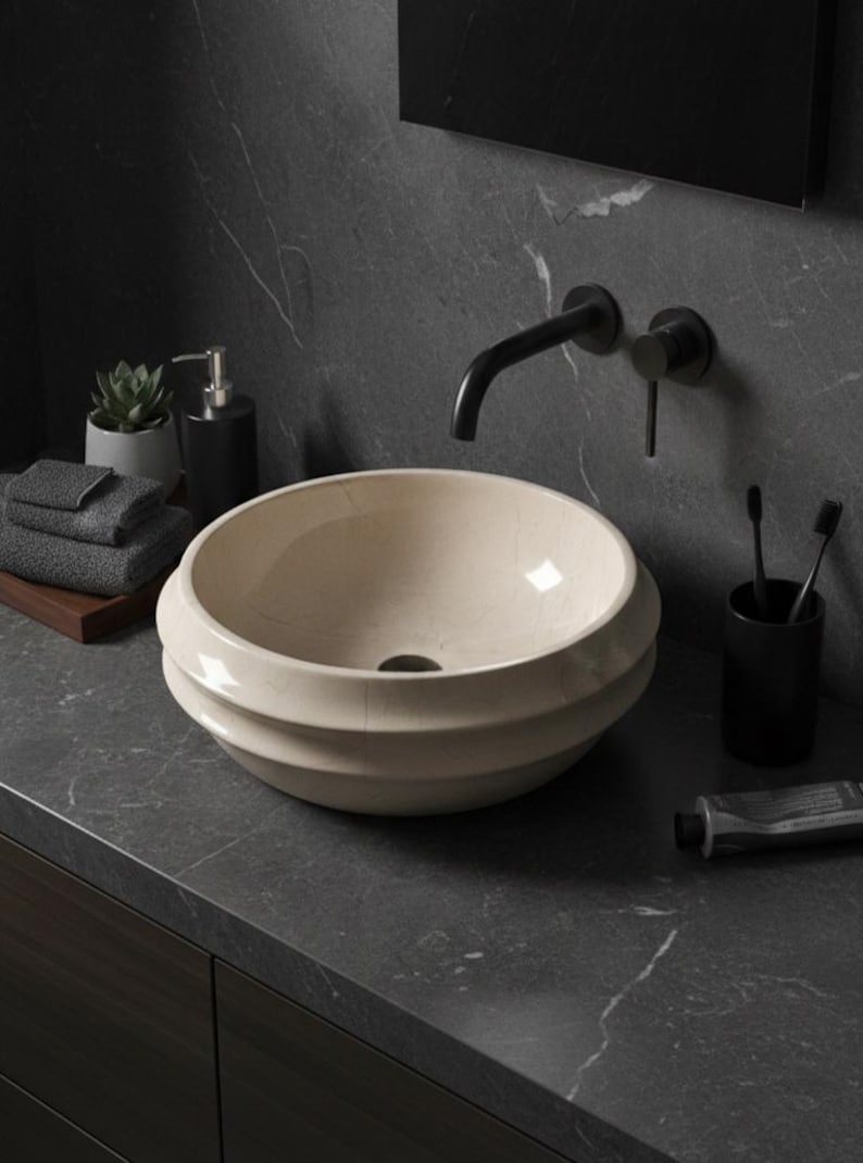 May include: A cream-colored, round bathroom sink with a tiered design. The sink is set on a dark gray countertop with a black faucet and a black soap dispenser. A small succulent, towels, and toiletries complete the modern bathroom scene.