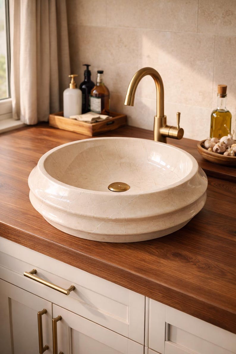 May include: A cream-colored, round bathroom sink with a decorative, tiered design. The sink has a gold-colored drain and is set on a dark wood countertop. A gold faucet is mounted above the sink. Other bathroom accessories are visible in the background.