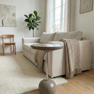 Pu&ograve; includere: Un moderno soggiorno con un divano beige, un tavolino rotondo con piano in marmo e una coperta intrecciata. La stanza ha una palette di colori neutri, una sedia in legno, un tappeto e una pianta.