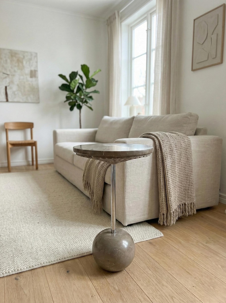 Pu&ograve; includere: Un moderno soggiorno con un divano beige, un tavolino rotondo con piano in marmo e una coperta intrecciata. La stanza ha una palette di colori neutri, una sedia in legno, un tappeto e una pianta.