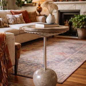 Pu&ograve; includere: Un tavolino decorativo con un piano rotondo e smerlato e un'unica gamba sottile color argento. La base ha una forma sferica. Un vaso bianco con fiori secchi e un libro sono appoggiati sul tavolo. Il tavolo &egrave; in un soggiorno.