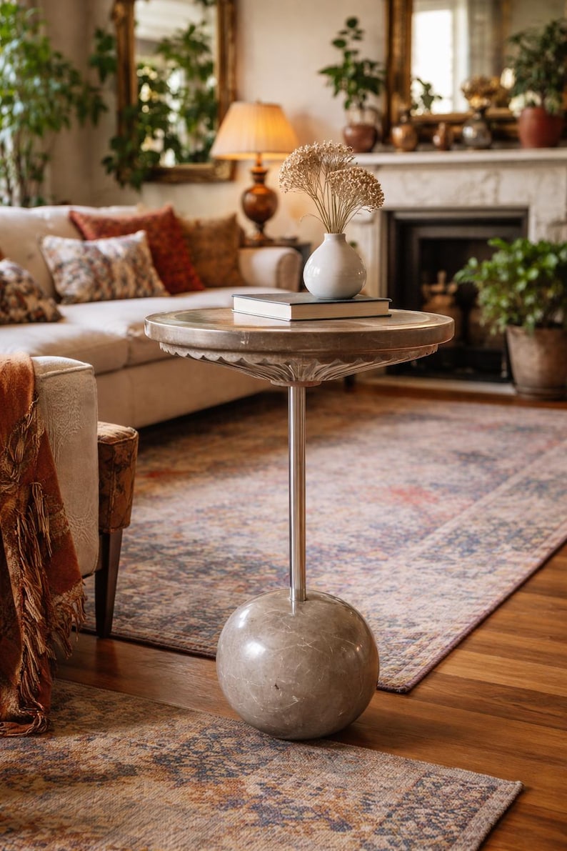 Pu&ograve; includere: Un tavolino decorativo con un piano rotondo e smerlato e un'unica gamba sottile color argento. La base ha una forma sferica. Un vaso bianco con fiori secchi e un libro sono appoggiati sul tavolo. Il tavolo &egrave; in un soggiorno.