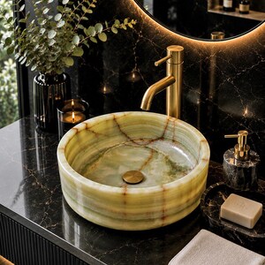 May include: A round vessel sink crafted from light green onyx stone with natural veining, featuring a brushed gold tap and drain. The sink sits on a black marble countertop next to a soap dispenser, a bar of soap, and a folded white towel.