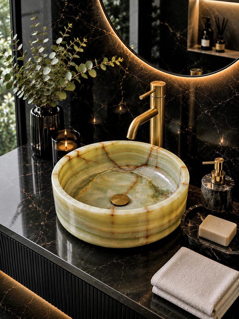May include: A round vessel sink crafted from light green onyx stone with natural veining, featuring a brushed gold tap and drain. The sink sits on a black marble countertop next to a soap dispenser, a bar of soap, and a folded white towel.