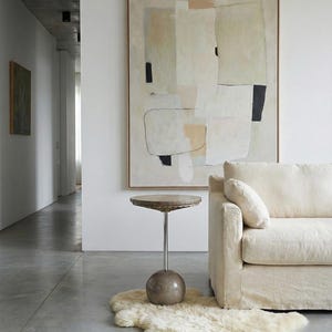 Pu&ograve; includere: Un dipinto astratto incorniciato &egrave; appeso sopra un divano color crema. Un piccolo tavolino rotondo con piano in pietra e base sferica poggia su un tappeto in pelle di pecora. La stanza ha un design minimalista con pavimento in cemento.