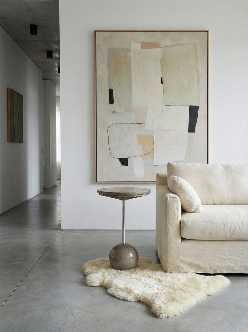 Pu&ograve; includere: Un dipinto astratto incorniciato &egrave; appeso sopra un divano color crema. Un piccolo tavolino rotondo con piano in pietra e base sferica poggia su un tappeto in pelle di pecora. La stanza ha un design minimalista con pavimento in cemento.