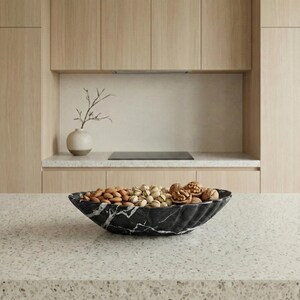 May include: A black marble serving bowl filled with almonds, pistachios, and walnuts. The bowl has a unique, wavy design and sits on a speckled countertop. The kitchen setting features light wood cabinets and a neutral color palette.