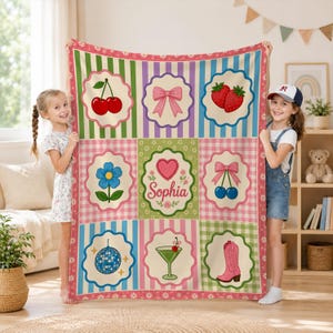 May include: A rectangular throw blanket featuring a grid of nine scalloped-edge panels on a striped and checkered background. The panels display illustrations of cherries, a pink bow, strawberries, a blue flower, a heart with the name Sophia, cherries with a bow, a disco ball, a martini glass, and a pink cowboy boot.