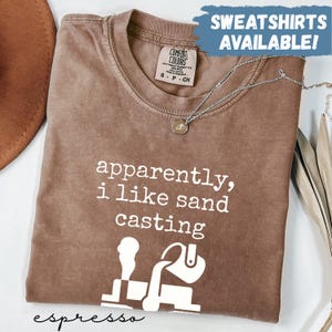 May include: Espresso-colored Comfort Colors t-shirt with the phrase "apparently, i like sand casting" in white. The shirt features a graphic of a sand casting process. A silver necklace with a pendant is draped on the shirt. The text "SWEATSHIRTS AVAILABLE!" is in the top right.