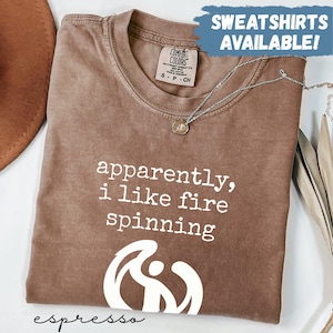 May include: Espresso-colored t-shirt with the text "apparently, i like fire spinning" in white. The shirt features a graphic of a person spinning fire and the words "SWEATSHIRTS AVAILABLE!" in a blue banner.