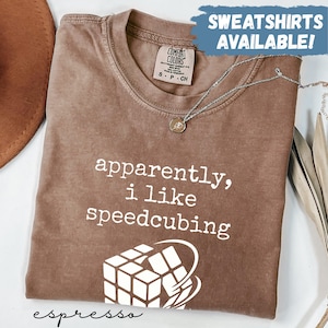 May include: Espresso-colored t-shirt with the phrase "apparently, i like speedcubing" in white. A graphic of a Rubik's Cube is below the text. The shirt has a Comfort Colors tag and a silver necklace.