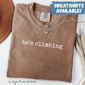 May include: Espresso-colored t-shirt with the phrase "he's climbing" in white lettering. The shirt has a crew neck and short sleeves. A silver necklace with a pendant is draped across the shirt. The words "SWEATSHIRTS AVAILABLE!" are in a blue banner.