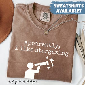 May include: Espresso-colored t-shirt with the phrase "apparently, i like stargazing" and a white graphic of a person looking through a telescope. The shirt has a crew neck and short sleeves. A silver necklace is draped across the shirt.