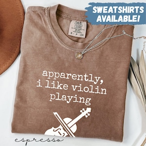 May include: Espresso-coloured t-shirt with the text "apparently, i like violin playing" and a violin graphic. The shirt has a crew neck and short sleeves. A silver necklace with a pendant is draped on the shirt. The words "SWEATSHIRTS AVAILABLE!" are in a blue box.