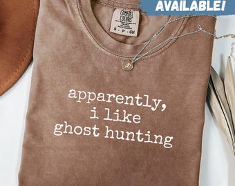 Könnte beinhalten: Braunes T-Shirt mit dem weißen Aufdruck "apparently, i like ghost hunting". Das Shirt hat einen Rundhalsausschnitt und ein Comfort Colors-Etikett. Eine silberne Halskette mit Anhänger ist über dem Shirt drapiert. Der Text "SWEATSHIRTS AVAILABLE!" steht oben rechts.