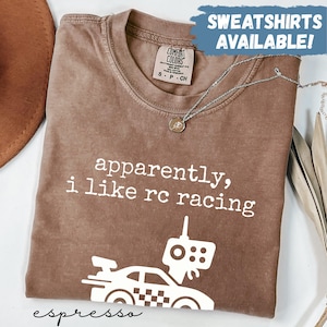 Pode incluir: Camiseta cor café com o texto "apparently, i like rc racing" e um gráfico de um carro de corrida e controle remoto. A camisa tem uma etiqueta "Comfort Colors". As palavras "Sweatshirts Available!" estão em uma caixa azul.