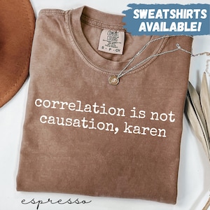 May include: A brown crew neck t-shirt with the white text "correlation is not causation, karen" printed on the front. The shirt features a Comfort Colors brand label at the inner neckline and a silver-toned chain necklace resting on the fabric.