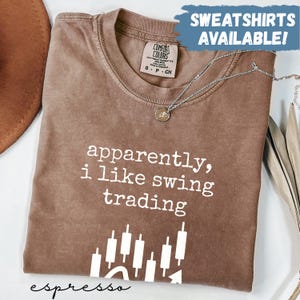 May include: Espresso-colored Comfort Colors sweatshirt with the text "apparently, i like swing trading" in white. The design includes a stock chart graphic. A silver necklace with a pendant is draped over the shirt. The words "SWEATSHIRTS AVAILABLE!" are in a blue box.