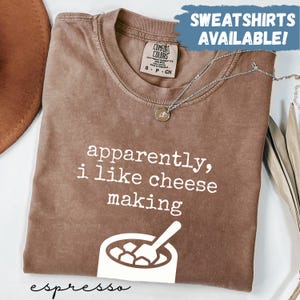 Op de afbeelding: Bruin t-shirt met de witte tekst "apparently, i like cheese making". Het shirt heeft een afbeelding van een kom met kaas en een lepel. De tekst "SWEATSHIRTS AVAILABLE!" staat in een blauwe doos.