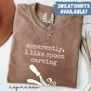 May include: Espresso-coloured Comfort Colors t-shirt with the text "apparently, i like spoon carving" in white. A silver necklace with a pendant is on the shirt. The words "SWEATSHIRTS AVAILABLE!" are in a blue box.
