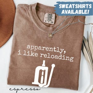 May include: Espresso-colored t-shirt with the text "apparently, i like reloading" and a graphic of a reloading tool. The shirt has a crew neck and short sleeves. A silver necklace with a pendant is draped across the shirt. The words "SWEATSHIRTS AVAILABLE!" are in the top right corner.