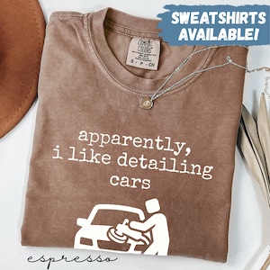 May include: Espresso-colored t-shirt with the text "apparently, i like detailing cars" and a graphic of a person detailing a car. The shirt also has the text "SWEATSHIRTS AVAILABLE!" and a Comfort Colors tag.
