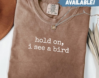 May include: Espresso-colored t-shirt with the text "hold on, I see a bird" in white. The shirt has a crew neck and a Comfort Colors tag. A silver necklace with a pendant is draped across the shirt. The words "SWEATSHIRTS AVAILABLE!" are in a blue box.