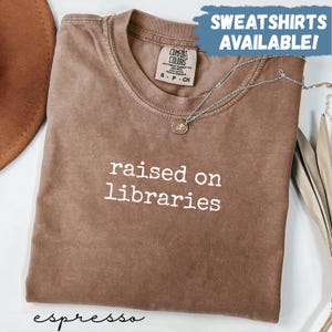 May include: A brown short-sleeved t-shirt with the white text "raised on libraries" printed on the chest. The shirt features a Comfort Colors brand label at the neck and a silver-toned necklace resting on the fabric. A blue graphic overlay reads "SWEATSHIRTS AVAILABLE!" and the word "espresso" is written in black script at the bottom.