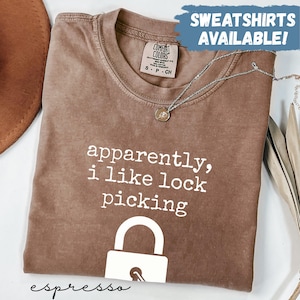 Puede incluir: Camiseta color espresso con el texto "apparently, i like lock picking" y un gráfico de candado. La camiseta tiene cuello redondo y mangas cortas. Un collar plateado con un colgante está drapeado sobre la camiseta. Las palabras "Sweatshirts Available!" están en un cuadro azul.