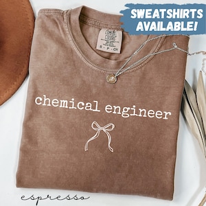 Puede incluir: Camiseta color espresso con las palabras "chemical engineer" en blanco y un gráfico de lazo. La camiseta tiene cuello redondo y mangas cortas. Un collar plateado con un colgante está drapeado sobre la camiseta. Las palabras "SWEATSHIRTS AVAILABLE!" están en un cuadro azul.