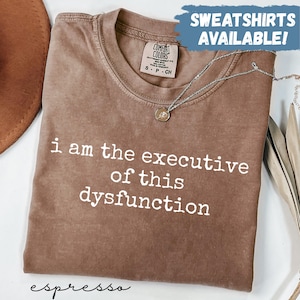Puede incluir: Camiseta color espresso con la frase "i am the executive of this dysfunction" en letras blancas. La camiseta tiene cuello redondo y mangas cortas. Un collar plateado con un colgante descansa sobre la camiseta. Las palabras "SWEATSHIRTS AVAILABLE!" están en un cuadro azul.