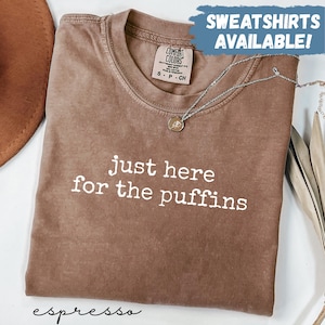 May include: A brown short-sleeve t-shirt with a crew neckline and the white text "just here for the puffins" printed on the front. The shirt features a Comfort Colors brand label at the inner neck and a blue graphic overlay stating "SWEATSHIRTS AVAILABLE!"