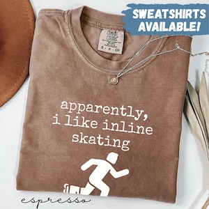 May include: Espresso-colored t-shirt with the text "apparently, i like inline skating" and a skater graphic. The shirt has a Comfort Colors tag and a silver necklace. The words "SWEATSHIRTS AVAILABLE!" are in a blue box.