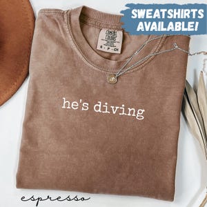 May include: Espresso-colored t-shirt with the phrase "he's diving" in white. The shirt is folded and has a Comfort Colors tag. A silver necklace with a pendant is draped across the shirt. Text at the top reads "SWEATSHIRTS AVAILABLE!"