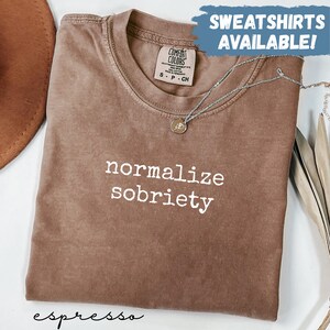 May include: Espresso-colored t-shirt with the phrase "normalize sobriety" in white. The shirt has a crew neck and a Comfort Colors tag. A silver necklace with a pendant is draped across the shirt. Text at the top says "SWEATSHIRTS AVAILABLE!"