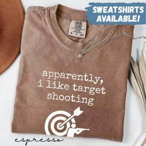 May include: Espresso-colored t-shirt with the text "apparently, i like target shooting" in white. A target and shooter graphic is below the text. The shirt has a Comfort Colors tag and a silver necklace.