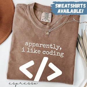 以下が含まれることがあります： 「apparently, i like coding」の白い文字とコード記号がプリントされた、エスプレッソ色のTシャツ。クルーネックで、Comfort Colorsのタグが付いています。シルバーのネックレスがTシャツの上にかけられています。「SWEATSHIRTS AVAILABLE!」の文字が背景にあります。