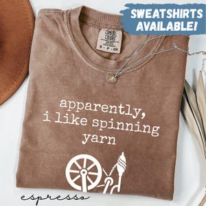 May include: Espresso-colored t-shirt with the text "apparently, i like spinning yarn" and a graphic of a spinning wheel and spindle. The shirt has a Comfort Colors tag and a silver necklace.