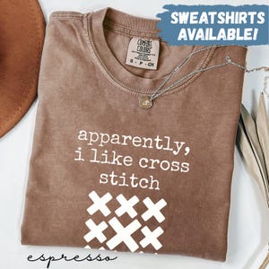 Può includere: T-shirt color espresso con la scritta "apparently, i like cross stitch" e grafica a punto croce. La maglietta ha girocollo e maniche corte. Una collana d'argento con un ciondolo è drappeggiata sulla maglietta. La scritta "SWEATSHIRTS AVAILABLE!" è sullo sfondo.