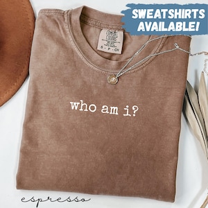 Puede incluir: Camiseta color espresso con la frase "who am i?" en letras blancas. La camiseta está doblada y tiene una etiqueta Comfort Colors. Un collar plateado con un colgante está sobre la camiseta. El texto superior dice "SWEATSHIRTS AVAILABLE!"