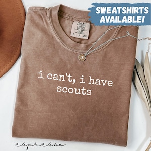May include: Espresso-colored t-shirt with the phrase "i can't, i have scouts" in white lettering. The shirt has a crew neck and a Comfort Colors tag. A silver necklace with a pendant is draped across the shirt. The words "SWEATSHIRTS AVAILABLE!" are in a blue banner.