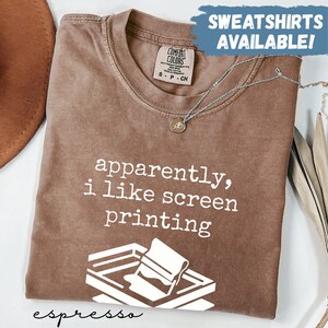 May include: Espresso-colored t-shirt with the text "apparently, i like screen printing" in white. The shirt features a screen printing graphic and the Comfort Colors brand tag. A silver necklace with a pendant is draped across the shirt.