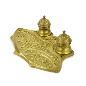 May include: Antique brass inkwell set with two ink pots and a decorative tray. The tray features floral and vine patterns. The ink pots have domed lids with ornate finials. The set is made of brass and has a gold finish.