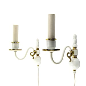 Può includere: Coppia di applique da parete in ceramica bianca con dettagli dorati. Ogni applique presenta un braccio curvo che sorregge un portacandele con una candela beige. Il design include dettagli decorativi e un cavo elettrico.