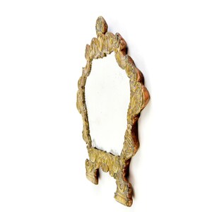 May include: Ornate, antique-style mirror with a decorative, gold-colored frame. The mirror has an oval shape and is supported by two small feet. The frame features intricate carvings and a detailed top design. The mirror is set against a white background.