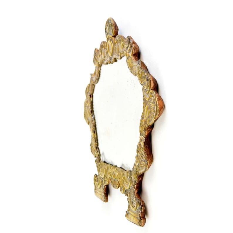 May include: Ornate, antique-style mirror with a decorative, gold-colored frame. The mirror has an oval shape and is supported by two small feet. The frame features intricate carvings and a detailed top design. The mirror is set against a white background.