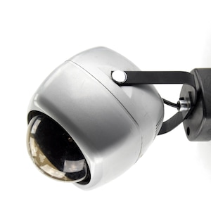 May include: A silver-colored security camera with a black mounting bracket. The camera has a rounded, dome-shaped lens and a metallic finish. The bracket is attached to a black base, suggesting a wall or ceiling mount.